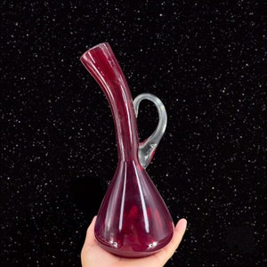 May include: A deep red glass decanter with a clear glass handle. The decanter has a long, curved neck and a round body.