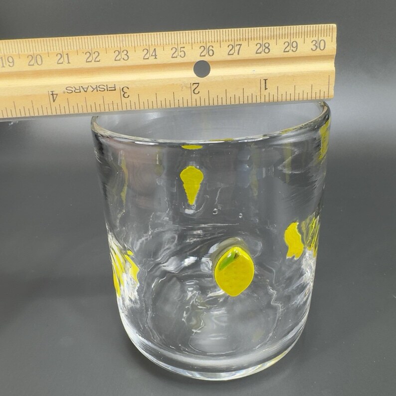May include: A clear glass tumbler with yellow abstract shapes. A wooden ruler is placed across the top of the glass, measuring approximately 3 inches in height. The glass has a rounded shape and is set against a dark background.