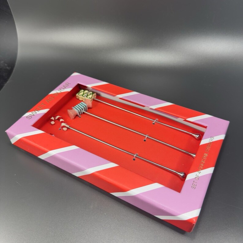 May include: A set of four stainless steel stir sticks with candy-themed toppers, presented in a red and pink striped box. The toppers include a candy cane, a wrapped candy, and a dessert with green frosting. The box reads "Sweet as Sugar".