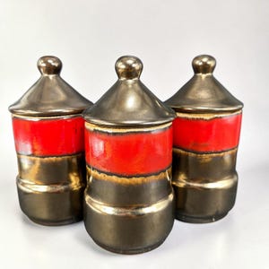 May include: Three cylindrical ceramic canisters with matching conical lids. Each canister features a metallic bronze finish on the base and lid, with a vibrant horizontal band of bright red glaze encircling the center of the body.