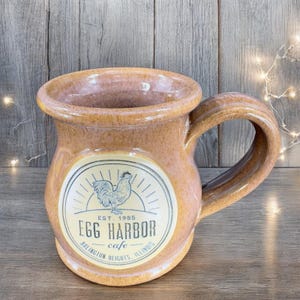 May include: A tan ceramic mug with a rooster and sun graphic and the text "Egg Harbor cafe Est. 1985 Arlington Heights, Illinois".