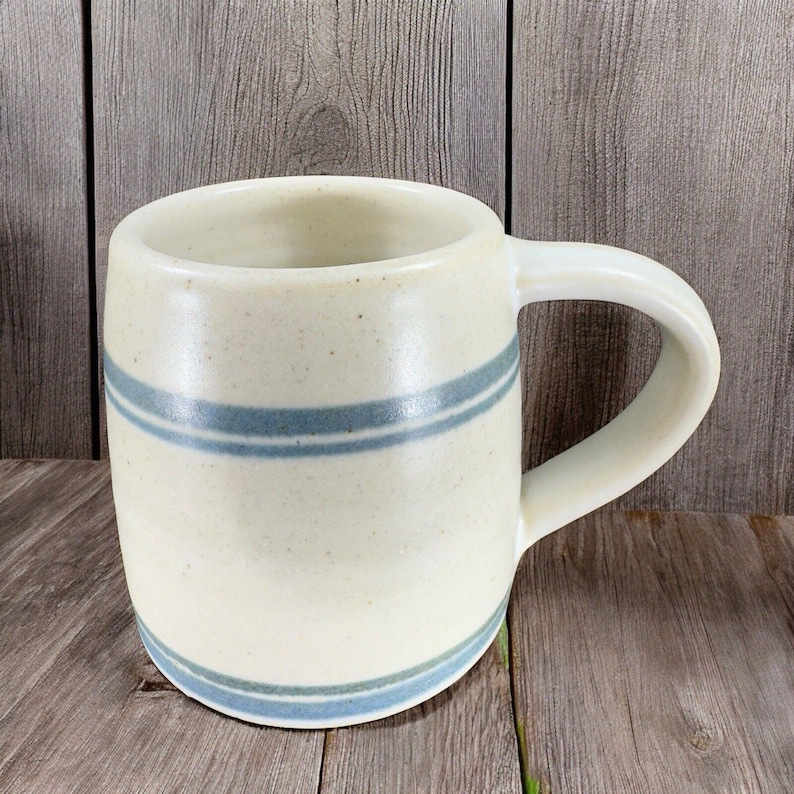 May include: Cream-colored ceramic mug with two horizontal blue stripes. The mug has a rounded body, a sturdy handle, and a speckled texture. The mug is sitting on a wooden surface.