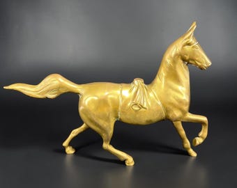 May include: A polished gold-toned brass horse figurine in a dynamic trotting pose. The sculpture features a smooth, reflective surface, a flowing tail, and a saddle detail on its back, captured in a side profile view with one front leg raised.