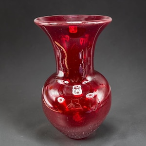 May include: A ruby red glass vase with a rounded body and flared rim. The vase has a smooth, glossy finish with small white circular accents. The vase is a decorative item, suitable for displaying flowers or as a standalone art piece.