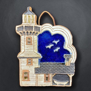May include: A ceramic wall hanging featuring a white lighthouse with a dark gray roof and a deep blue sky background. The piece includes a small brown leather hanging loop at the top and depicts three white birds flying in the blue sky area.