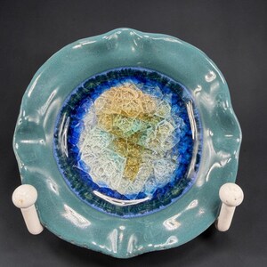 May include: A teal ceramic ashtray with a ruffled edge. The center features a mosaic design in shades of blue, yellow, and white. Two white, mushroom-shaped holders are attached to the sides. The ashtray is a decorative item.