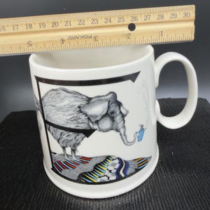 May include: White ceramic mug with a black and white elephant illustration. The elephant is holding a blue watering can. The mug has a black border and a colorful patterned base. The ruler shows the mug is approximately 4 inches tall.
