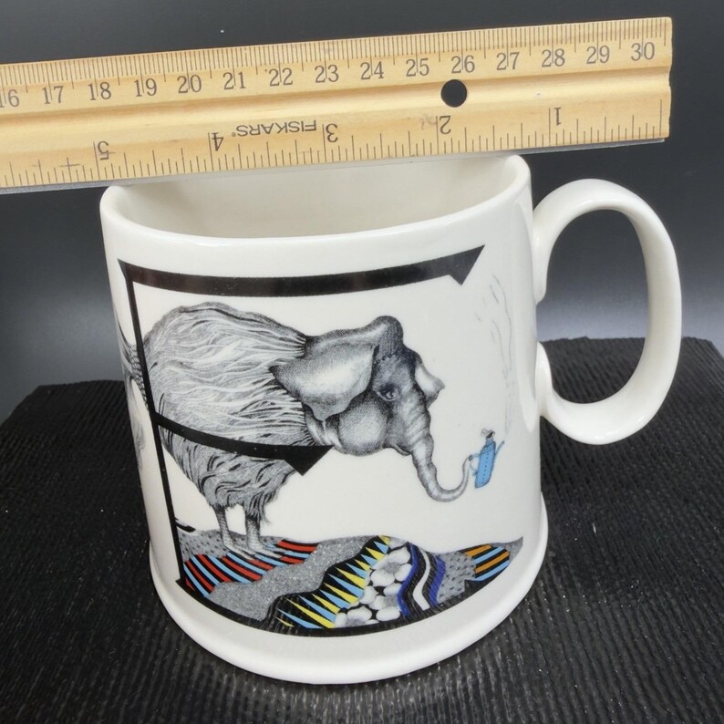 May include: White ceramic mug with a black and white elephant illustration. The elephant is holding a blue watering can. The mug has a black border and a colorful patterned base. The ruler shows the mug is approximately 4 inches tall.