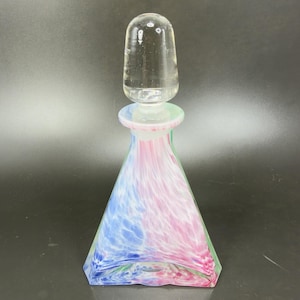 May include: A decorative glass bottle with a clear stopper. The bottle has a flared, geometric shape and is swirled with blue, pink, and green colors. The stopper is clear and rounded. The bottle is likely for perfume or other liquids.