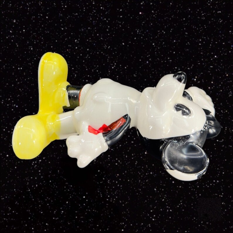 K&ouml;nnte beinhalten: Eine wei&szlig;e Keramikfigur von Mickey Mouse, die auf dem R&uuml;cken liegt, mit gelben Schuhen, schwarzen Ohren und einer roten Schleife.