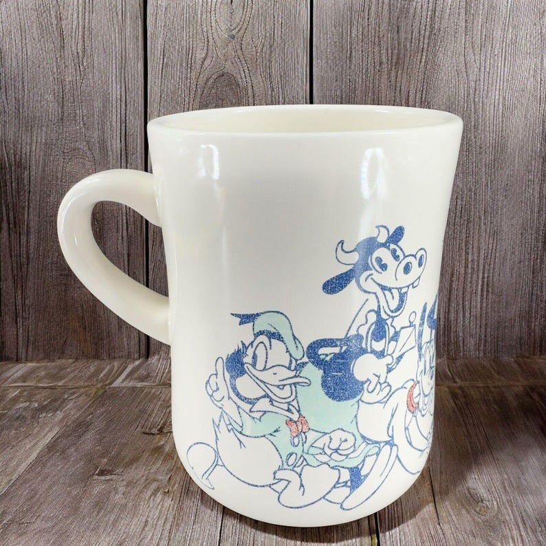 May include: White ceramic mug with a blue and green cartoon design featuring Mickey Mouse, Donald Duck, Goofy, and Pluto.