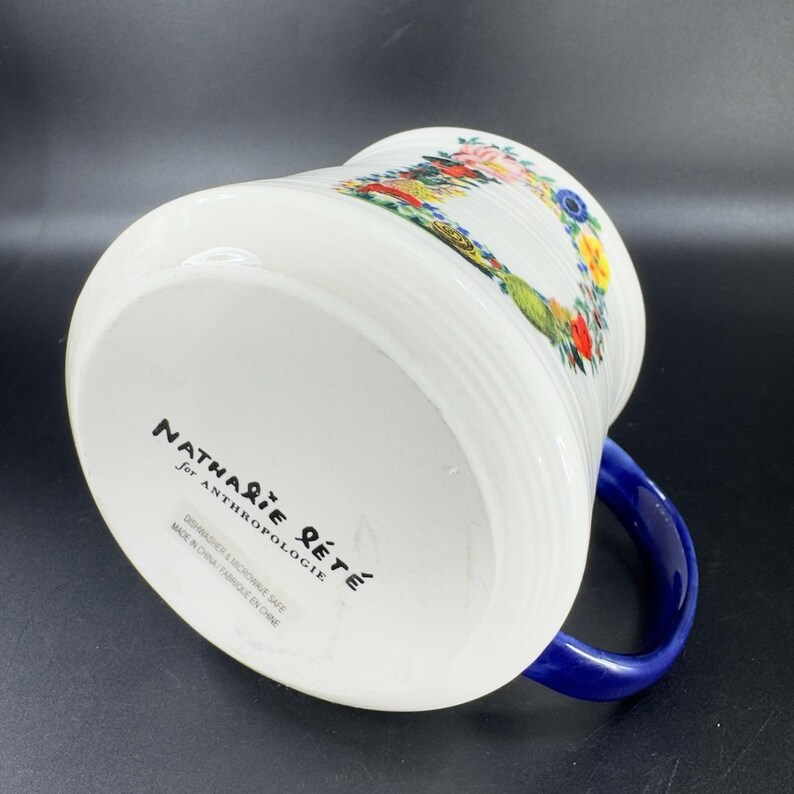 Puede incluir: Taza de cer&aacute;mica blanca con asa azul, con un dise&ntilde;o floral y de p&aacute;jaros. La taza tiene detalles acanalados verticales y el texto "Nathalie Lete for Anthropologie" est&aacute; impreso en la parte inferior.