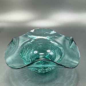 May include: A teal-colored glass bowl with a ruffled rim. The bowl has a crackle finish, giving it a unique texture. The bowl's design is reminiscent of a flower, with the ruffled edges adding a decorative touch. The bowl is likely used for serving or display.