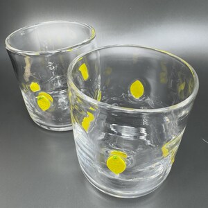May include: Two clear, round drinking glasses with a yellow lemon design. The glasses are made of clear glass and have a slightly textured surface. The lemons are painted in a bright yellow color. The glasses are set against a dark gray background.