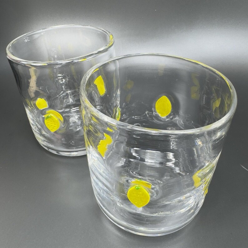 May include: Two clear, round drinking glasses with a yellow lemon design. The glasses are made of clear glass and have a slightly textured surface. The lemons are painted in a bright yellow color. The glasses are set against a dark gray background.
