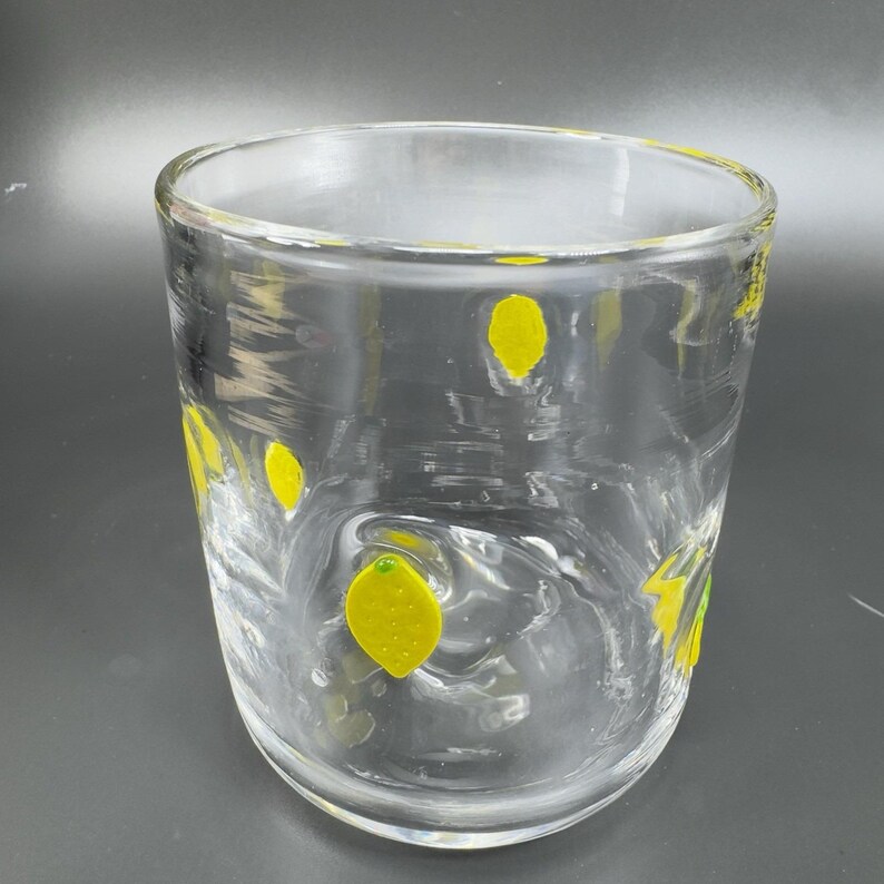 May include: A clear glass tumbler decorated with yellow lemon designs. The glass has a wide mouth and a slightly curved body. The lemons are evenly spaced around the glass, adding a touch of whimsy to the design.
