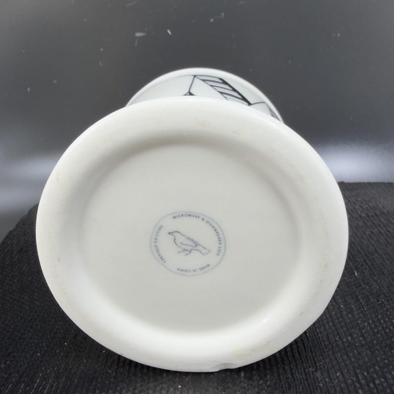 May include: White ceramic mug with a black graphic design and a bird logo on the bottom. The mug is labeled "Microwave & Dishwasher Safe" and "Made in China".