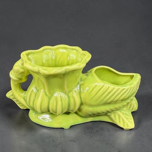 May include: A lime green ceramic planter with two sections. One section is a cup-shaped opening, and the other is a boat-shaped opening. The planter has a floral design and is set on a base.