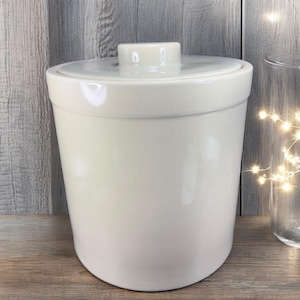 May include: A cream-colored ceramic container with a lid and a small knob. The container is cylindrical with a slightly rounded base. The lid fits snugly on top. The background is a wooden surface.