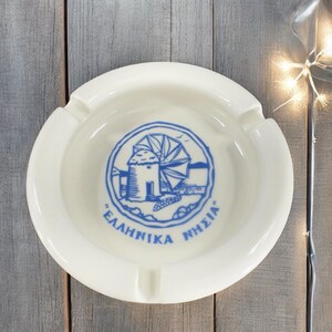 May include: White ceramic ashtray with a blue and white windmill design. The design includes the text "ΕΛΛΗΝΙΚΑ ΝΗΣΙΑ".