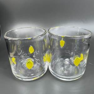 May include: Two clear, round drinking glasses with yellow lemon designs. The glasses are made of clear glass and have a slightly textured appearance. The yellow lemon designs are painted on the outside of the glasses.
