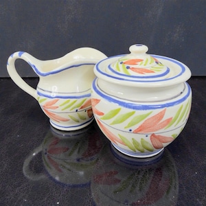 May include: A cream-colored ceramic creamer and sugar bowl set. The set features a blue rim and is decorated with red, green, and blue leaf and floral patterns. The sugar bowl has a matching lid.