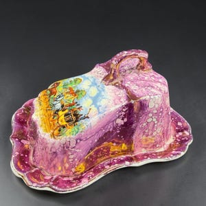 May include: A ceramic butter dish with a purple and white marbled glaze finish. The lid features a colorful illustration of a horse-drawn carriage with the text "Coaching Days" and has a small handle on top. The base has a scalloped edge.