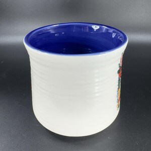 Puede incluir: Porta utensilios de cer&aacute;mica blanca con interior azul y dise&ntilde;o floral. El exterior tiene una textura acanalada. El dise&ntilde;o floral incluye colores rojo, amarillo y verde. El soporte es cil&iacute;ndrico y tiene una abertura ancha.