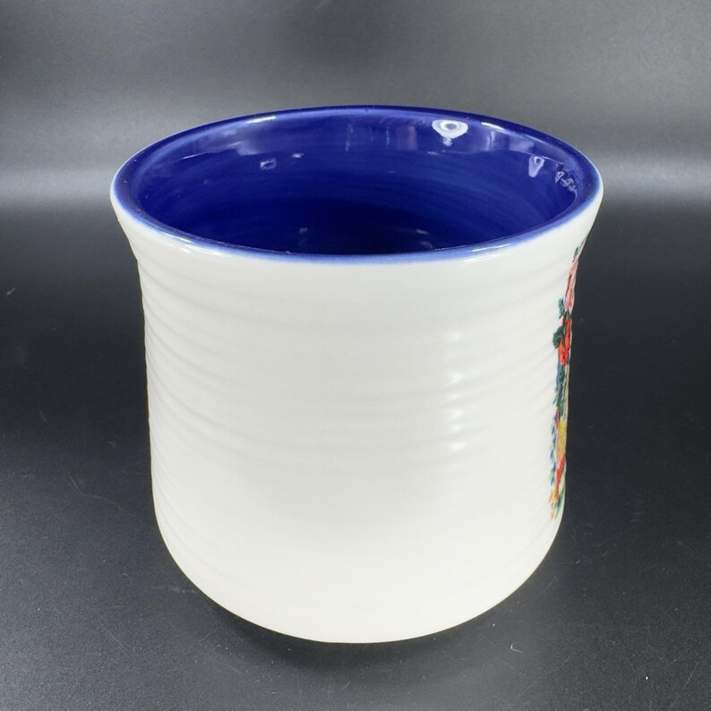 Puede incluir: Porta utensilios de cer&aacute;mica blanca con interior azul y dise&ntilde;o floral. El exterior tiene una textura acanalada. El dise&ntilde;o floral incluye colores rojo, amarillo y verde. El soporte es cil&iacute;ndrico y tiene una abertura ancha.