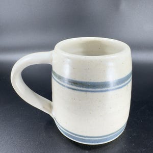 May include: Cream-colored ceramic mug with a rounded body and a white handle. The mug features two horizontal blue stripes around the middle. The mug is designed for hot beverages.