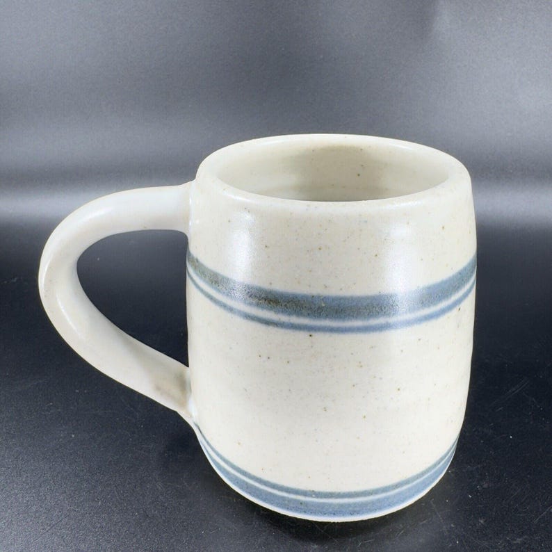 May include: Cream-colored ceramic mug with a rounded body and a white handle. The mug features two horizontal blue stripes around the middle. The mug is designed for hot beverages.