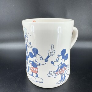 May include: White ceramic mug with a blue and red design featuring Mickey Mouse, Minnie Mouse, and Goofy. The characters are in a cartoon style and are smiling.
