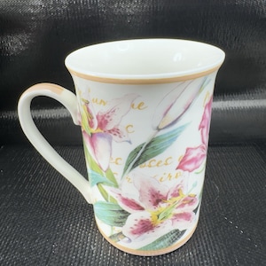 May include: White ceramic mug with a floral design featuring pink and white lilies, green leaves, and gold script. The mug has a curved handle and a gold rim. The background is black.