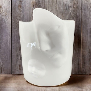 May include: White ceramic vase with an abstract, organic shape. The vase has a smooth, glossy finish and a unique, flowing design.