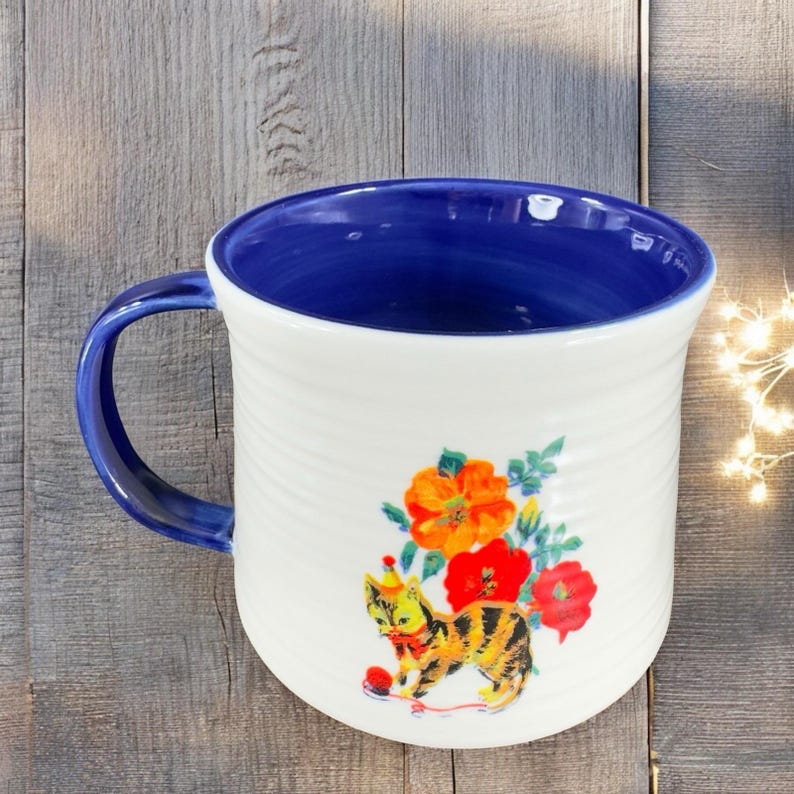Puede incluir: Taza de cer&aacute;mica blanca con interior y asa azules. La taza presenta una colorida ilustraci&oacute;n de un gato con gorro de fiesta y flores. La taza tiene una textura acanalada.