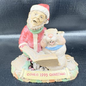 May include: A Christmas figurine featuring a man in a red Santa hat and coat, reading a book with a rabbit. The figurine has a beige base with the text "COIR 1995 CHRISTMAS". The man has a white beard and green scarf.