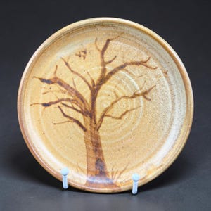 May include: A round, tan-colored ceramic plate with a hand-painted brown tree design. The tree's trunk and branches are depicted in detail, with a textured surface. The plate is a decorative piece for serving or display.
