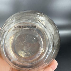 May include: Clear glass bottle with a textured surface. The bottom of the bottle has a label that reads "by ANTHROPOLOGIE".