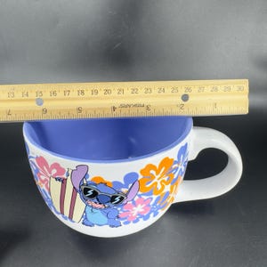 May include: White ceramic mug with a blue, orange, and pink floral design. The mug features a cartoon image of Stitch from Lilo & Stitch wearing sunglasses and holding a surfboard. The mug has a blue interior and a handle.