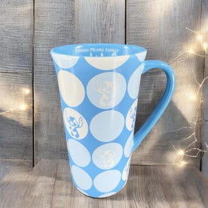 May include: A blue ceramic mug with a white polka dot pattern and a white silhouette of Stitch from Lilo & Stitch. The mug has a handle and says "Ohana Means Family" on the side.