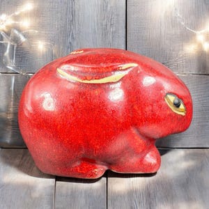 May include: A glossy red ceramic bunny figurine with yellow accents on the ears and around the eye. The bunny is in a seated position, with a smooth, rounded body. The background is a wooden surface.