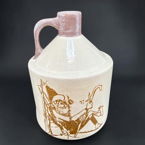 May include: A cream-colored ceramic jug with a glossy finish and a light brown handle. The body features a brown graphic illustration of a bearded character sitting in a chair, holding a pipe and a tool, with a rifle leaning against the chair.