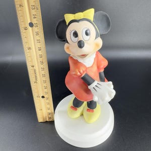 May include: A porcelain figurine of Minnie Mouse wearing a red dress with a yellow bow in her hair. She is standing on a white base.
