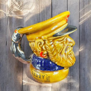 May include: A vintage ceramic mug shaped like a man's head, featuring a yellow hat and beard, a blue scarf, and the name "Barry" on a banner. The handle is silver. The mug is on a wooden surface.