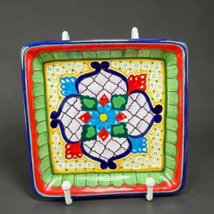 May include: Square, hand-painted ceramic plate with a vibrant floral design. The plate features a central flower with blue, red, and green petals, surrounded by a white and yellow patterned background. The rim is bordered with red, green, and blue.
