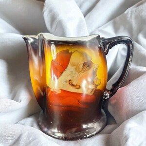 May include: A brown and gold ceramic creamer with a painted image of a man wearing a red hat and robe. The creamer has a handle and a spout.