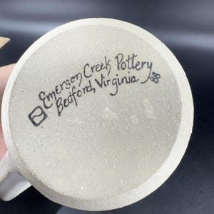 May include: The bottom of a ceramic mug with the text "Emerson Creek Pottery, Bedford, Virginia" written in black ink. The mug is off-white with a speckled texture.