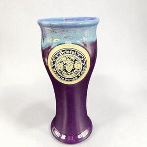 May include: A tall, flared ceramic pilsner glass featuring a deep purple glaze with a light blue rim. A circular cream-colored medallion is centered on the front, displaying a jester illustration and the text Bristol Renaissance Faire 2005.