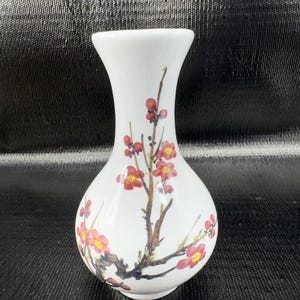 May include: A white ceramic vase with a slender neck and rounded body. The vase is decorated with a hand-painted design of red and yellow flowers on brown branches. The vase is a decorative item.