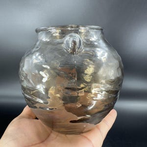 May include: A clear glass vase with a textured surface and a handle on the side. The vase has a rounded shape and a slightly iridescent finish.